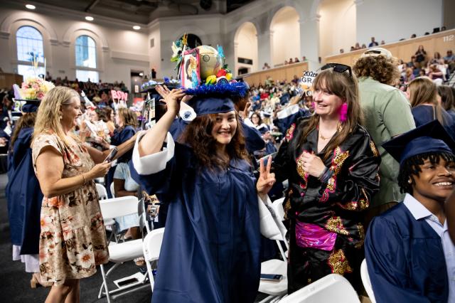 Somaya Ahmadi ’26 is capped during Convocation by Juanita Giles - A graduate in a navy gown smiles and flashes a peace sign in an auditorium. The mortarboard is decorated with a globe and bright blue trim. Guests and other graduates stand nearby taking photos and chatting. Crowded seating and arched walls frame the ceremony in the background.