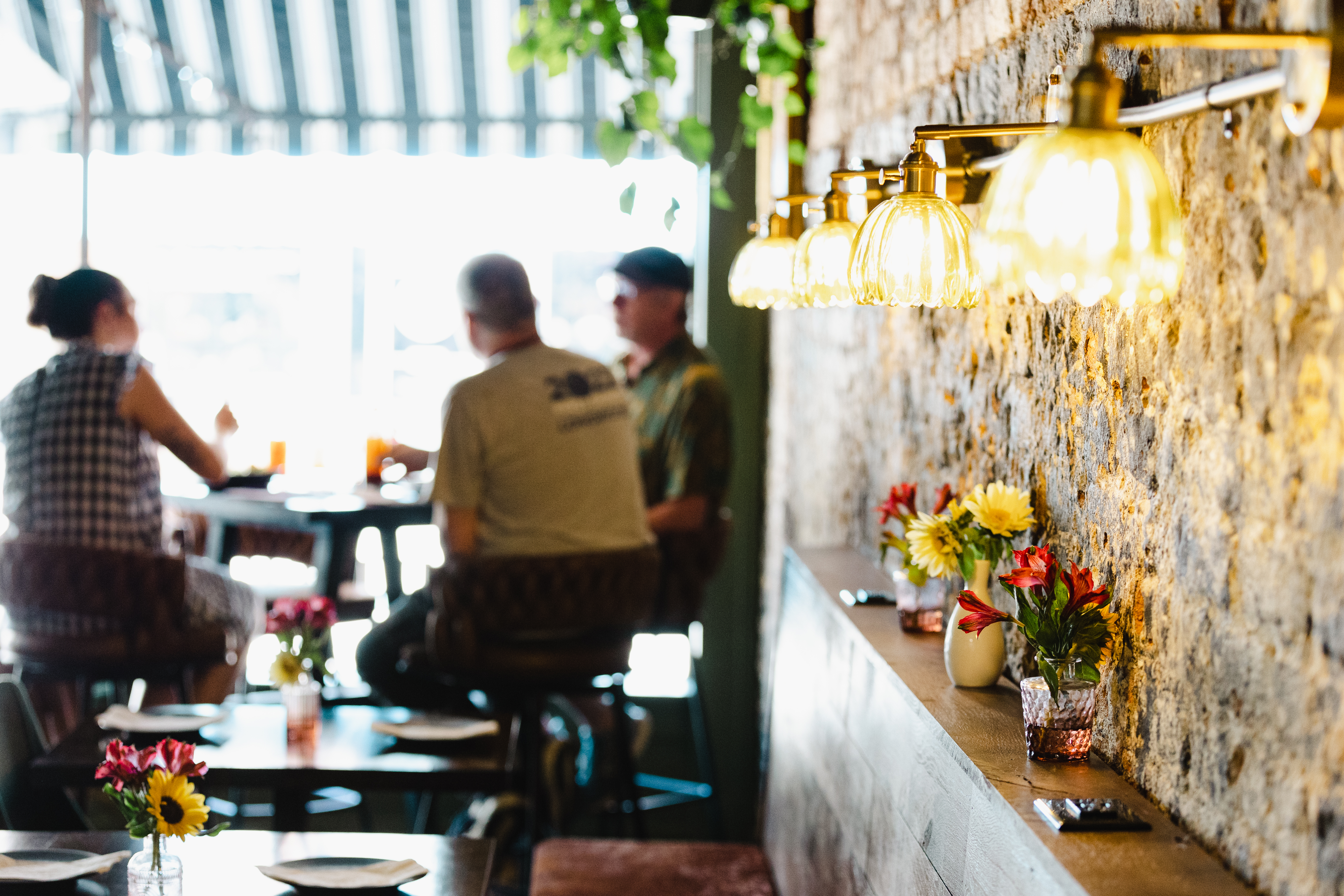 Diners inside The Fox and the Lamb - Warm pendant lights glow along a brick wall inside a restaurant. Small vases of colorful flowers line a narrow ledge beside the wall. Diners sit at tables near a bright front window with a striped awning. The scene is softly focused, emphasizing the cozy interior ambiance.