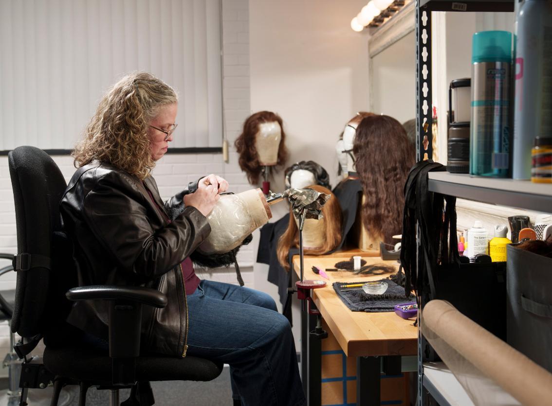 Samantha Wootten '00 works at a table, styling a wig on a mannequin head. She wears glasses and a black leather jacket, focused on her hands as she adjusts the piece. Several wigs on mannequin heads line the work surface and background. Hair tools, products, and supplies are spread across the workspace under a lighted mirror.