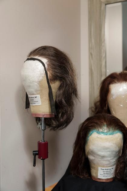 Two wigs are displayed on mannequin heads in Samantha Wootten's workshop. One wig is mounted on a stand, while another sits on a table beside it. Labels on the mannequin heads identify the performers and productions. A mirror and additional wigs appear softly in the background.