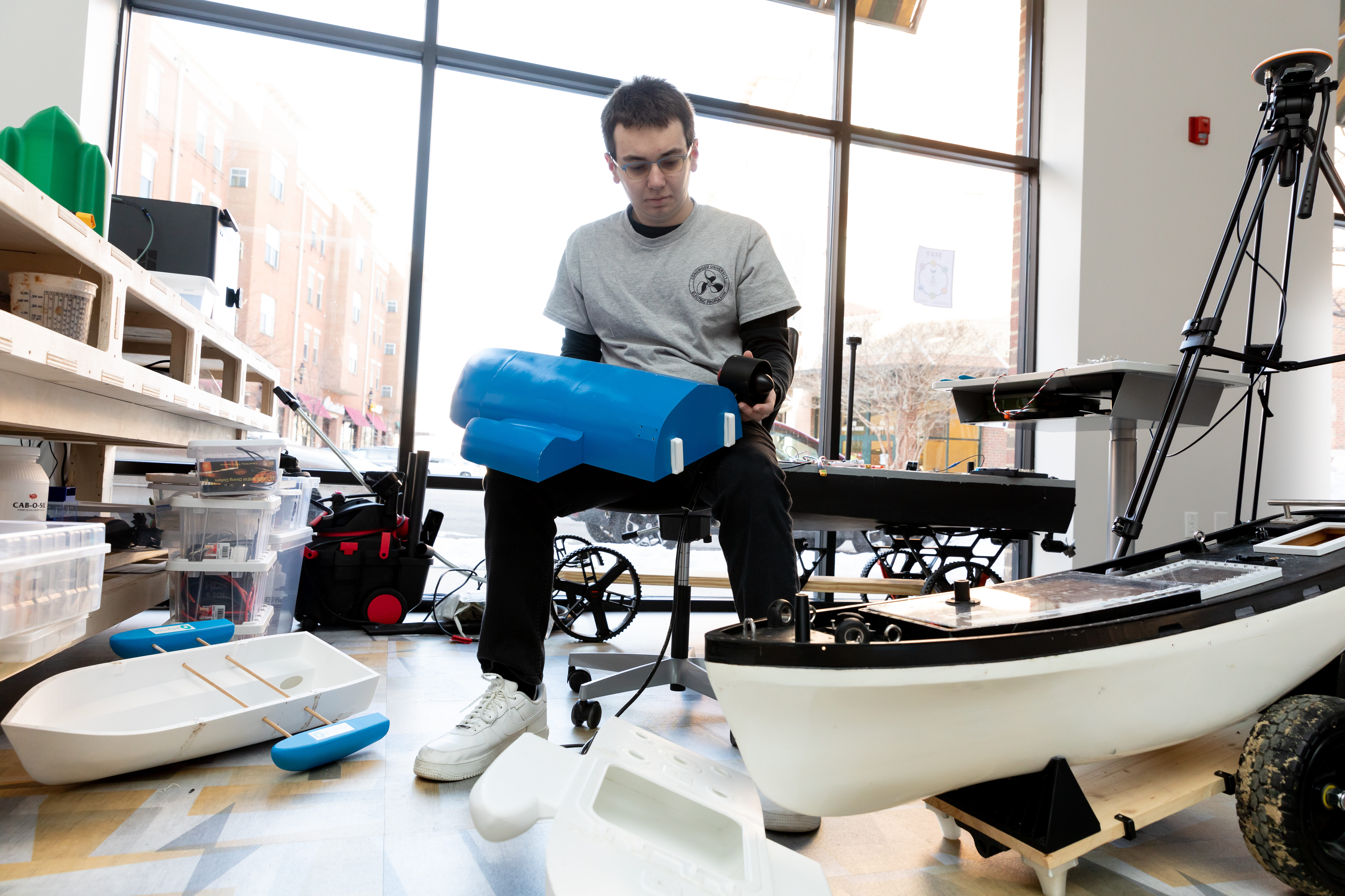 Senior Ben Gettier works on a portion of his nautical device in Longwood's SEED Innovation Hub. He is sitting in front of a window, on a rolling chair, holding two parts to the device.