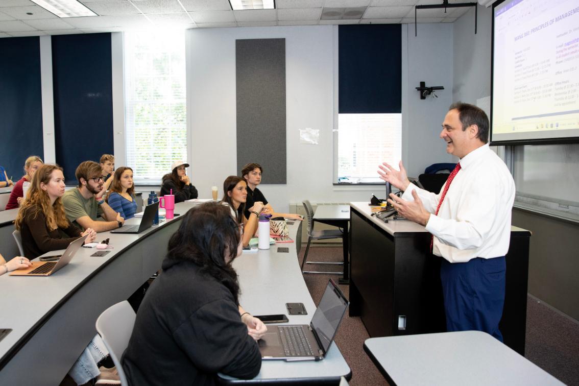 Dr. Vince Magnini lectures at the front of a classroom, gesturing as he speaks. Students sit in tiered rows, listening and taking notes on laptops. A projected screen displays course material beside him. Large windows and acoustic panels line the bright classroom.