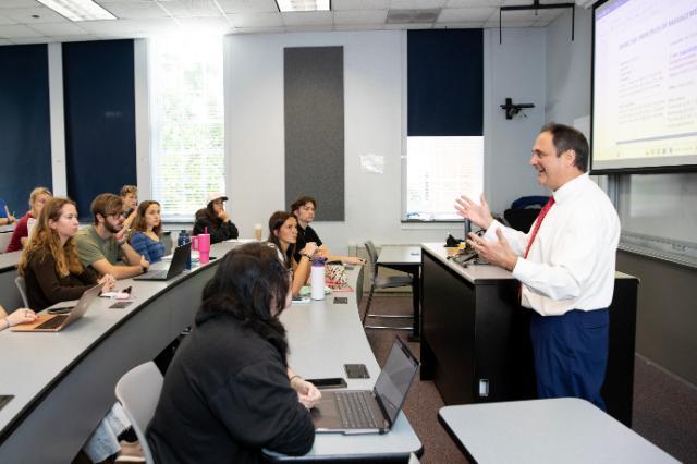 Dr. Vince Magnini lectures at the front of a classroom, gesturing as he speaks. Students sit in tiered rows, listening and taking notes on laptops. A projected screen displays course material beside him. Large windows and acoustic panels line the bright classroom.