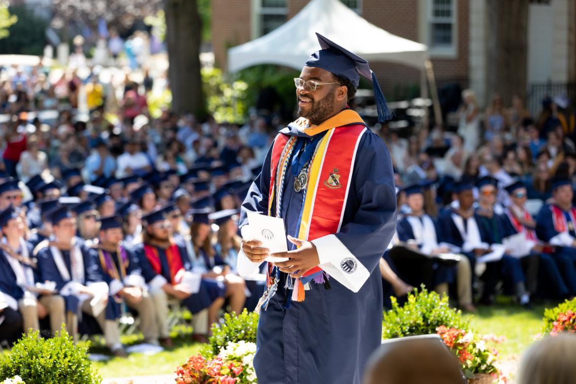 A Longwood University undergraduate graduate smiles while walking during Commencement, wearing a blue cap and gown with honor cords and a red stole as classmates look on from Wheeler Mall.