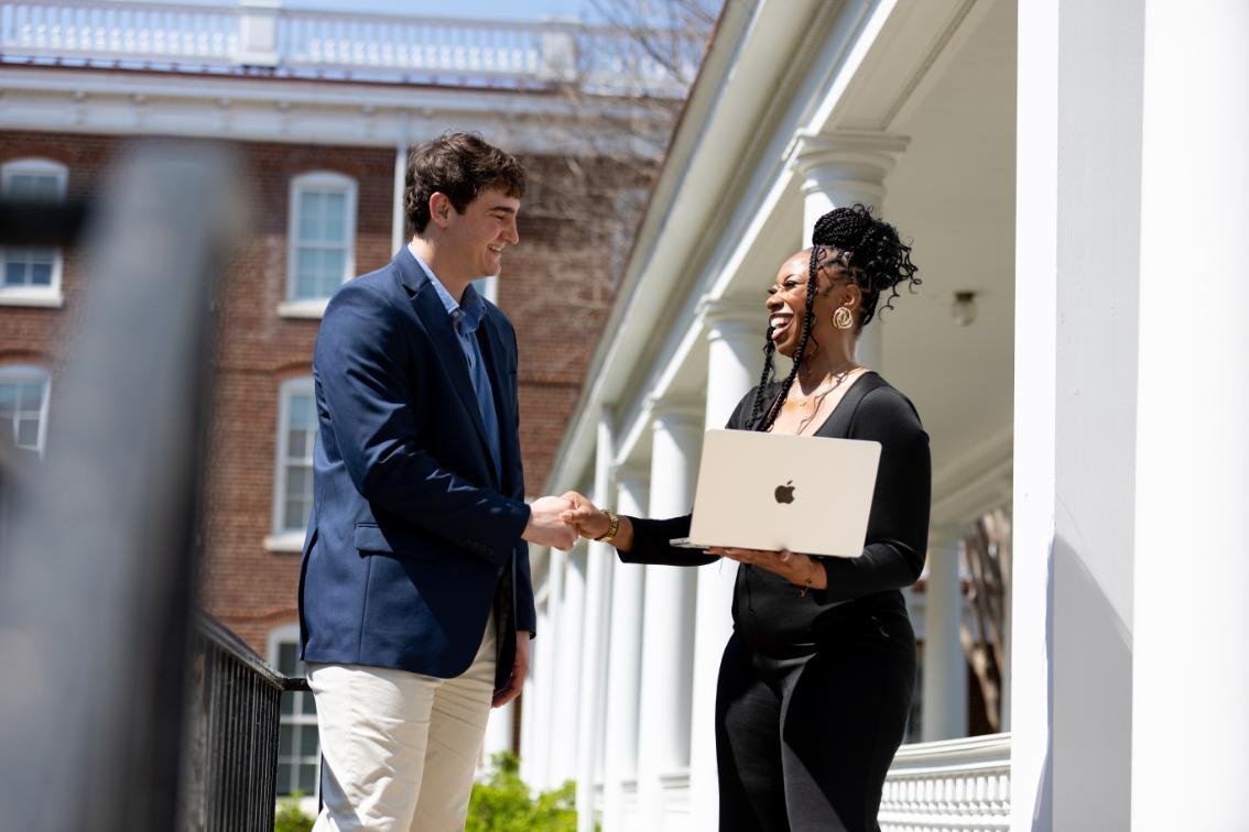 Two Master of Science in Information Technology students laugh and shake hands outside a Longwood building, one holding a laptop as they stand beside white columns.