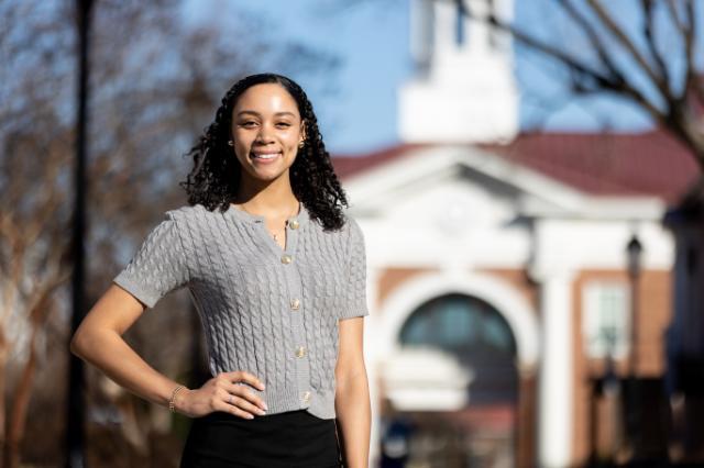Photo of Naiya Casimiro standing confidently with her right hand on her hip, and with Allen Hall in the background.