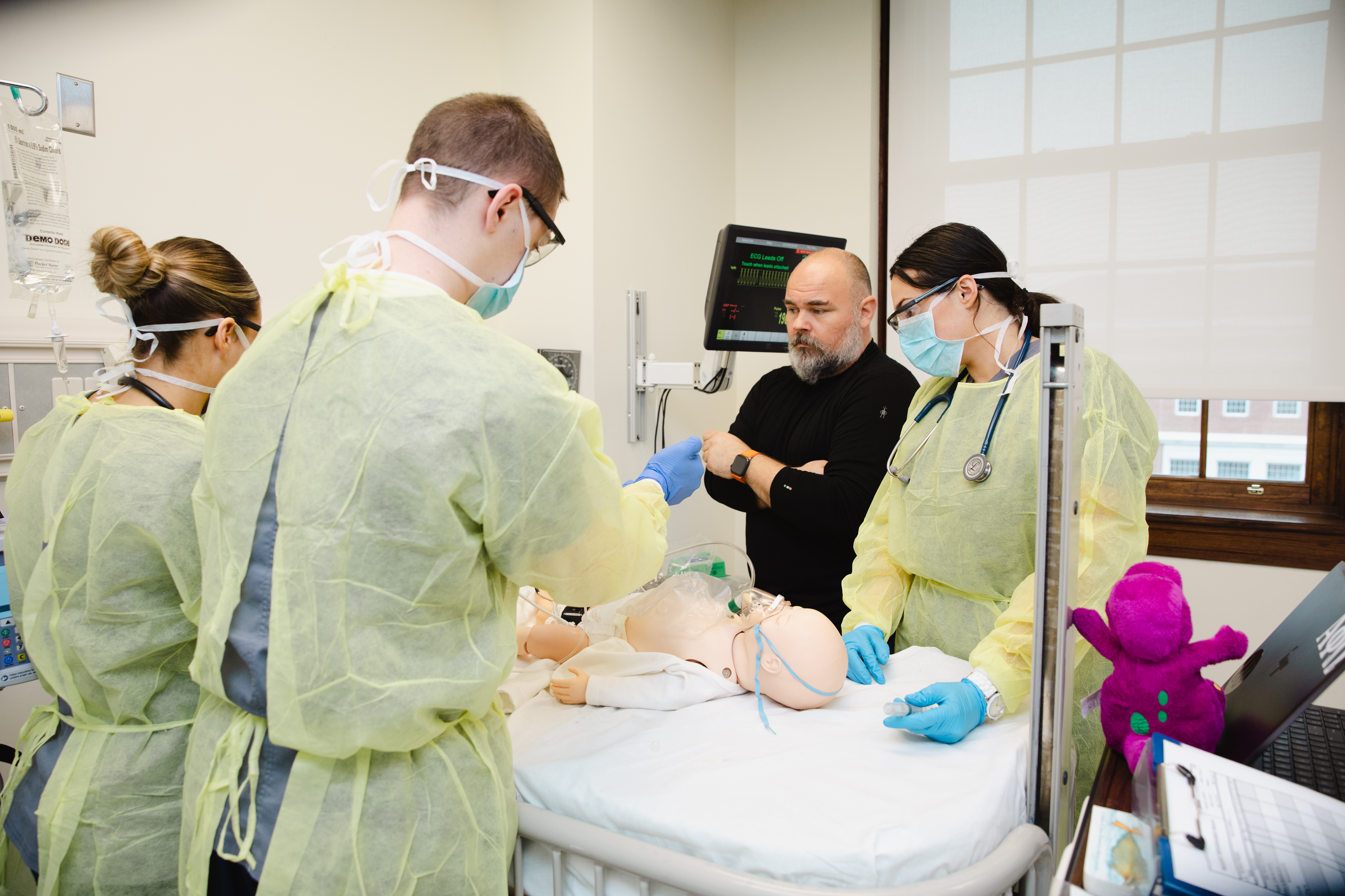 Longwood nursing students in protective gowns and masks practice patient care with a simulation mannequin as a faculty member observes in the nursing simulation lab.