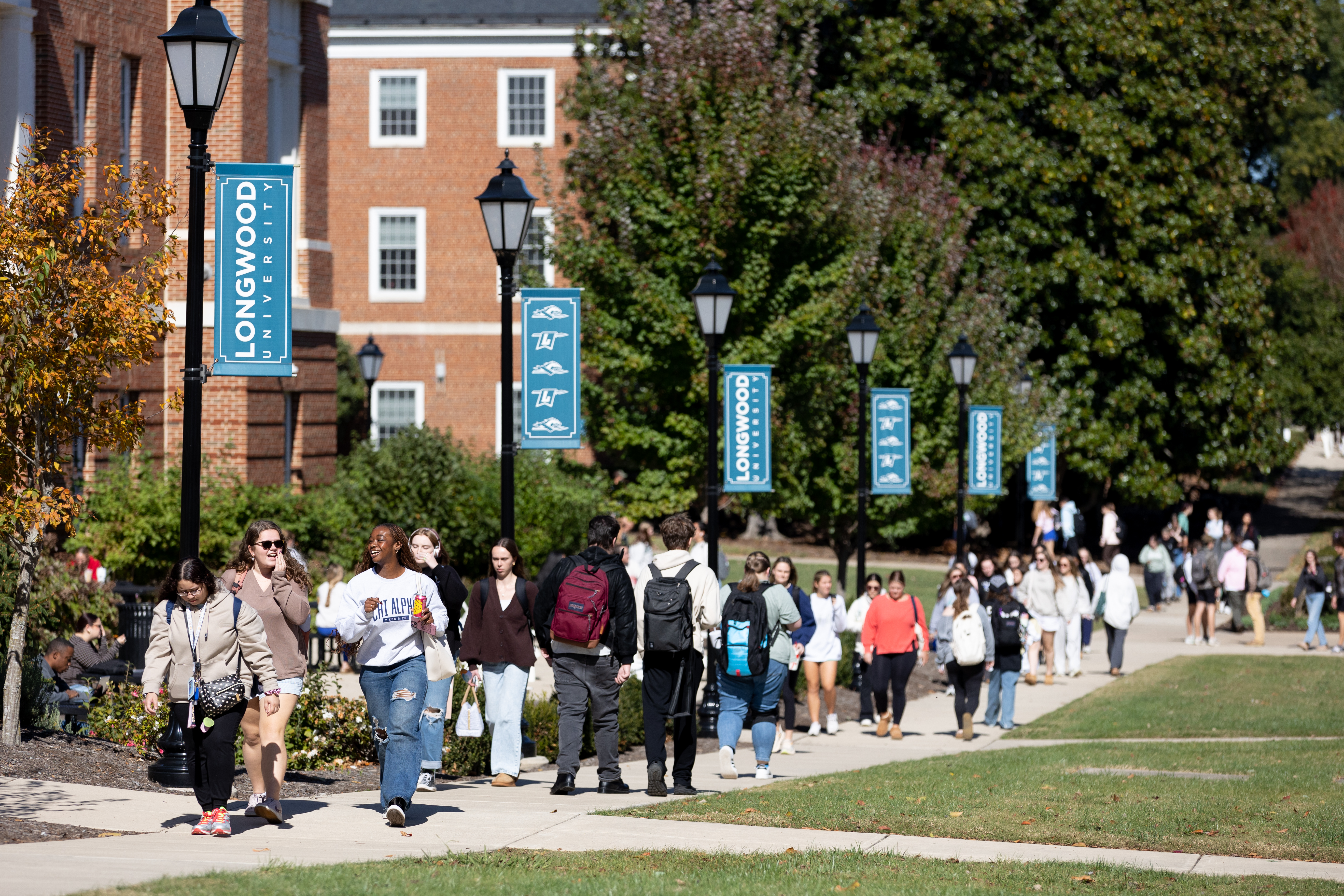 Students walk and talk along a busy campus sidewalk lined with Longwood banners on a sunny fall day.