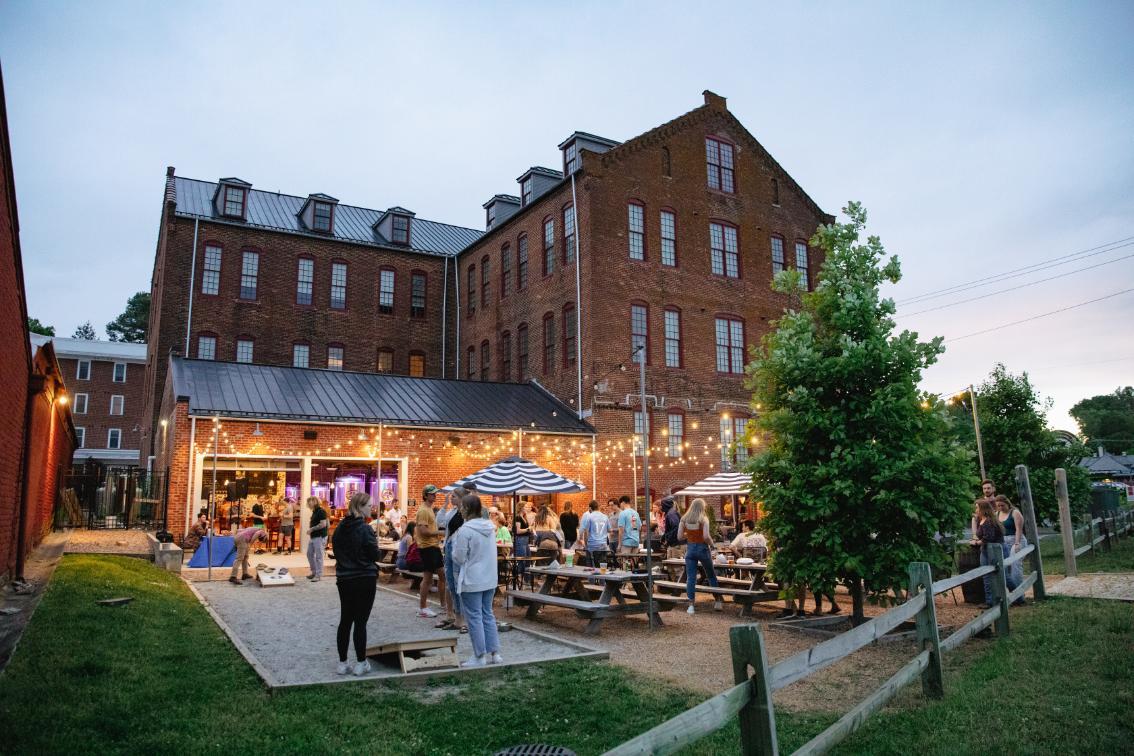 Students and friends hanging out at Three Roads Brewing during Senior Week - Outdoor gathering at dusk beside a tall brick building with many windows. String lights hang over picnic tables and umbrellas on a patio area. People mingle and play games in the courtyard near an open bar entrance. A wooden fence and small trees frame the scene under an evening sky.
