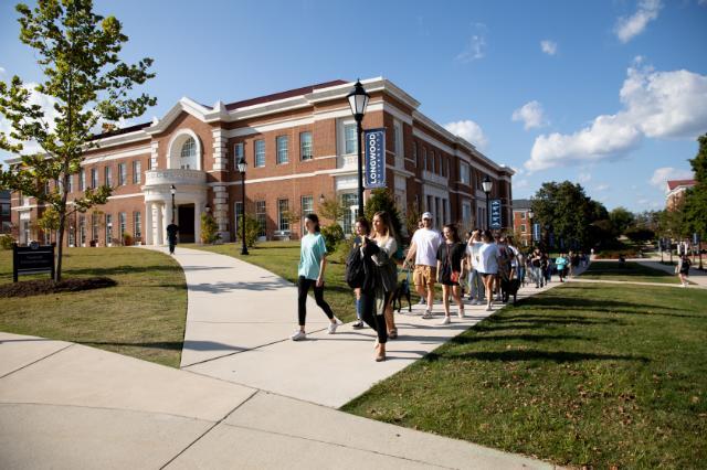 Students walk together past Longwood’s Upchurch University Center on a sunny day, following a campus sidewalk lined with Longwood banners.