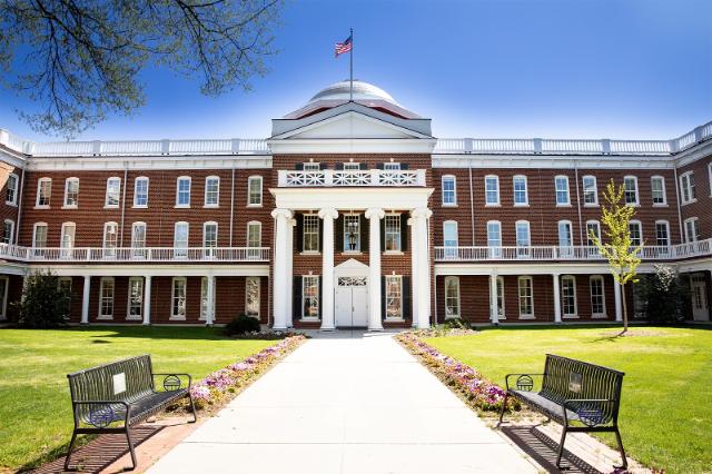 Rotunda Hall - Front view of a red-brick campus building with white columns and a domed roof. An American flag flies above the central entrance under a clear blue sky. A wide walkway leads to the doors, bordered by flower beds and green lawns. Two benches sit symmetrically along the path in the foreground.