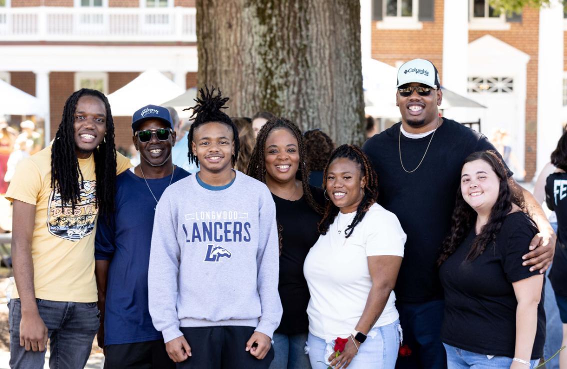 A family smiles together during Longwood Family Weekend, with campus tents and visitors gathered behind them.