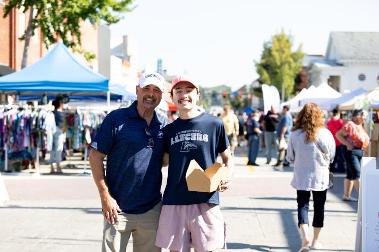 Two people smile together in Longwood gear during Family Weekend, with vendors and campus visitors in the background.